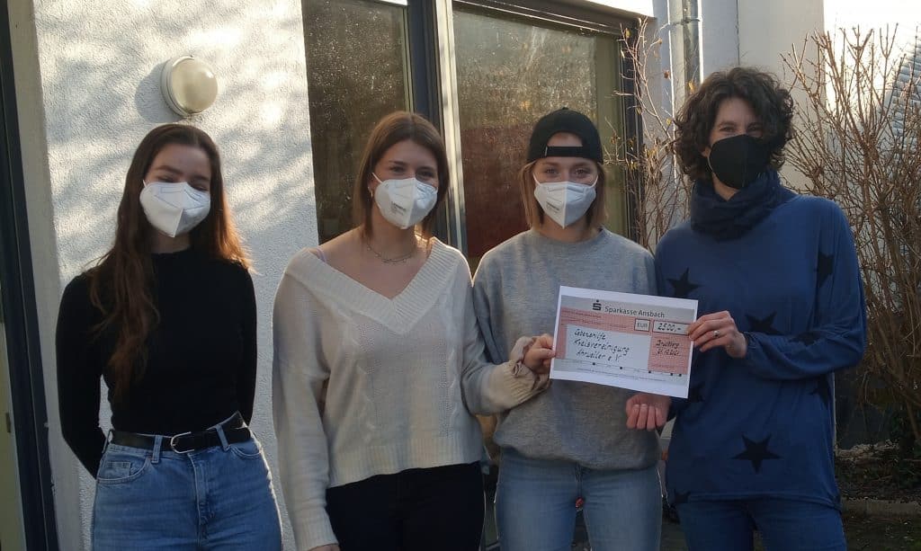 Vier maskierte Personen stehen draußen, zwei halten gemeinsam eine Urkunde oder einen Scheck.
