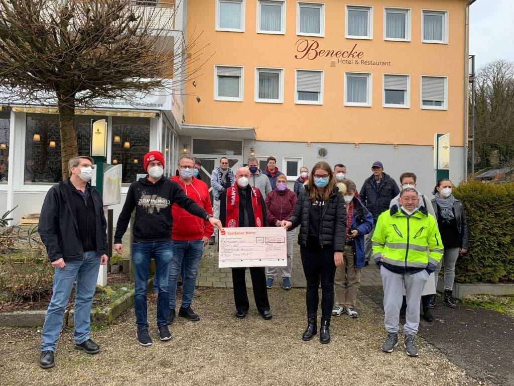 Eine Gruppe maskierter Personen hält einen großen Scheck vor einem Hotel-Restaurant.