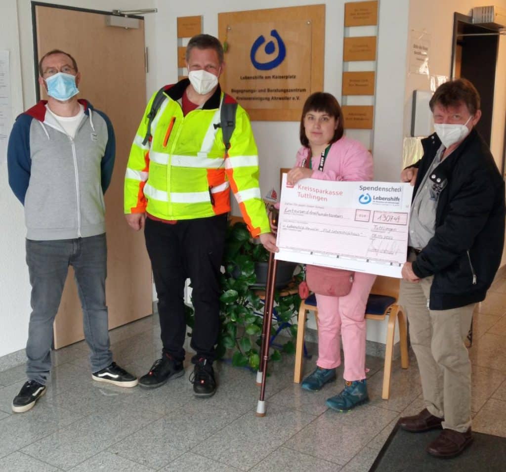 Vier Personen, drei davon mit Masken, halten einen großen Spendenscheck in der Nähe eines Wandschildes.