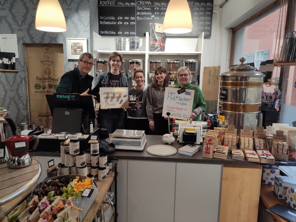 Fünf Personen stehen hinter einem Tresen in einem Café, halten Schilder und lächeln in die Kamera.