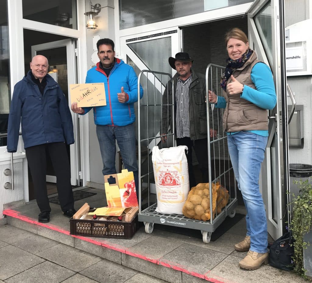 Vier Personen stehen neben einem Wagen mit Lebensmitteln vor einem Gebäudeeingang und posieren für ein Foto.