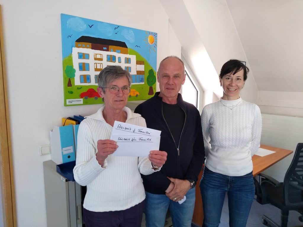 Drei Menschen stehen in einem Haus, einer hält ein Papier mit handgeschriebenem Text in der Hand, in der Nähe eines bunten Gemäldes.