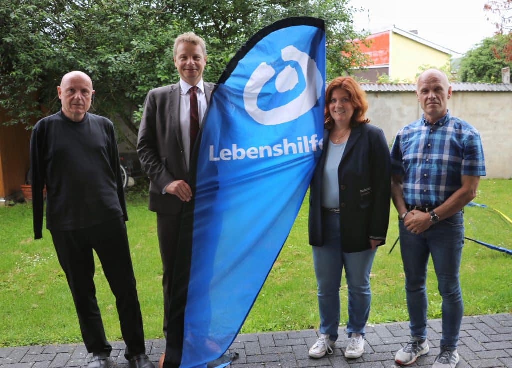 Vier Erwachsene stehen im Freien neben einer blauen Fahne mit der Aufschrift "Lebenshilfe".