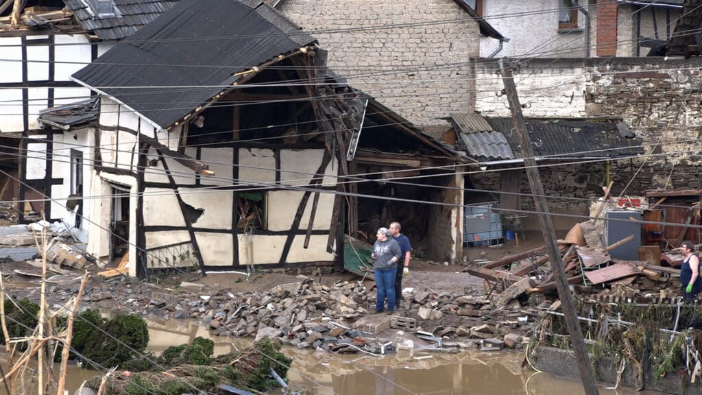 Zwei Menschen stehen inmitten von Hochwassertrümmern vor einem beschädigten Haus mit eingestürzten Wänden und Dach.
