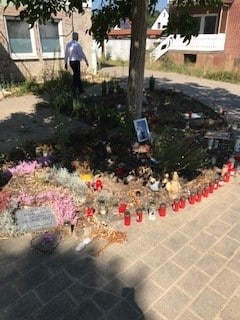 Eine kleine Gedenkstätte mit Blumen, Kerzen und Fotos neben einem Baum an einem sonnigen Tag.