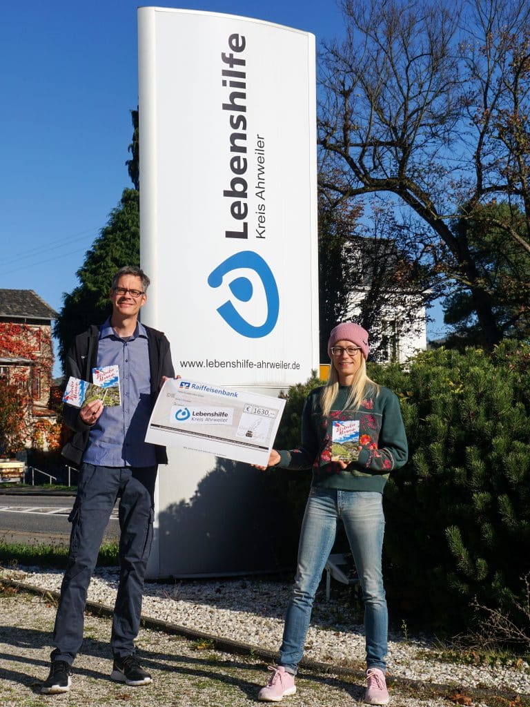 Zwei Personen stehen draußen und halten einen großen Scheck vor einem Schild der Lebenshilfe Ahrweiler.