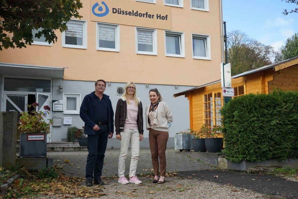 Drei Personen stehen vor dem Gebäude des Düsseldorfer Hofs auf einer gepflasterten Fläche in der Nähe einer Grünanlage.
