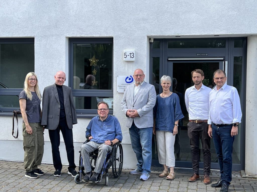 Sieben Erwachsene, darunter ein Rollstuhlfahrer, stehen vor einem Gebäude mit der Aufschrift 5-13.