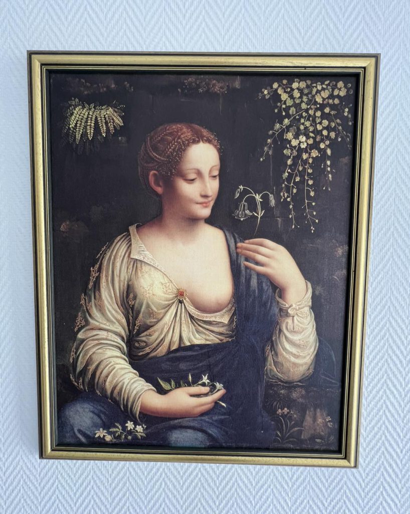Ein gerahmtes Gemälde einer Frau in Renaissancekleidung, die Pflanzen hält, hängt an einer strukturierten Wand.