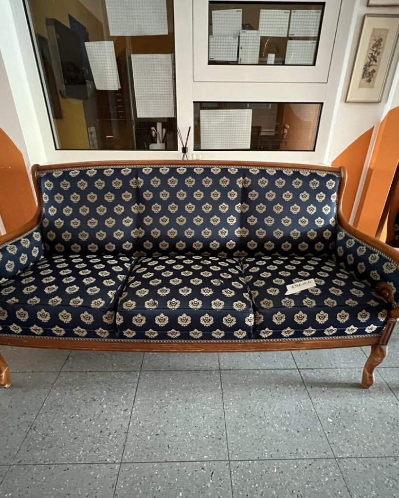 Ein dunkelblau gemustertes Sofa mit Holzrahmen, das auf einem gefliesten Boden vor den Fenstern steht.