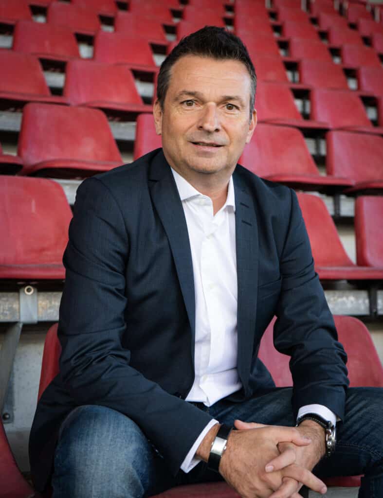Ein Mann im Anzug sitzt auf roten Stadionsitzen, die Hände gefaltet, und schaut in die Kamera.