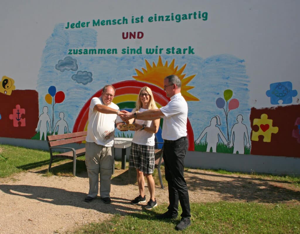 Drei Menschen stehen lächelnd vor einem bunten Wandgemälde mit deutschen Texten und gemalten Figuren.