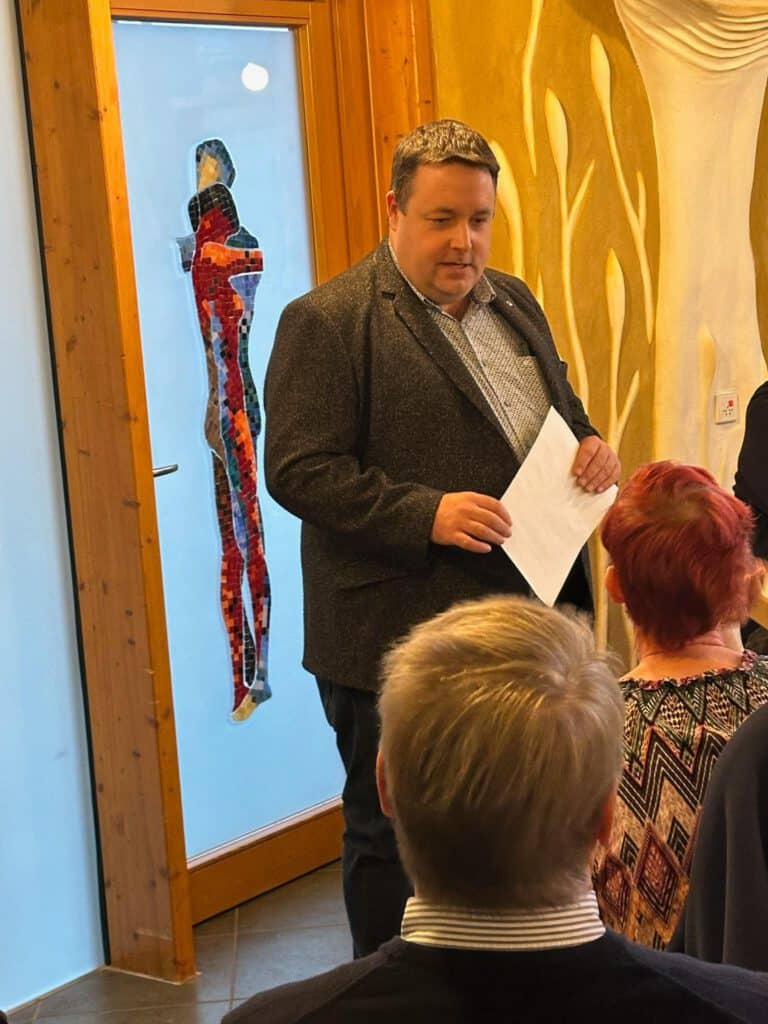 Ein Mann, der Papiere in der Hand hält, spricht zu einem sitzenden Publikum in der Nähe einer Tür mit einem farbenfrohen menschlichen Figurenmuster.