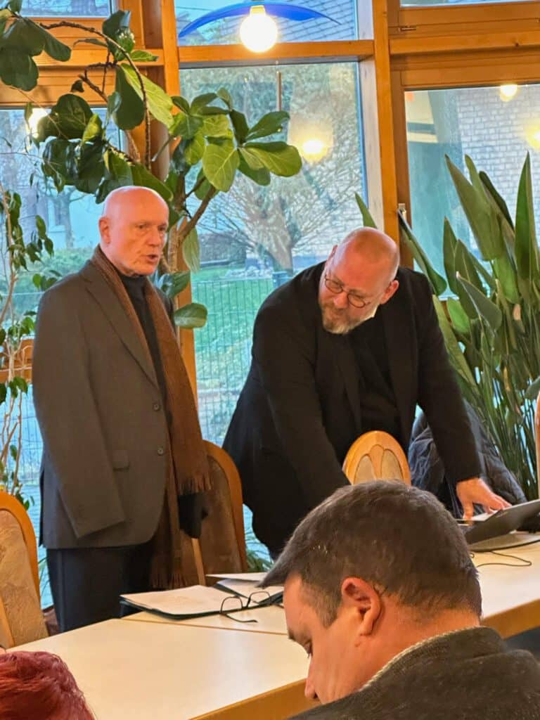 Zwei Männer in Anzügen stehen an einem Tisch in einem gut beleuchteten Raum mit großen Pflanzen und unterhalten sich.