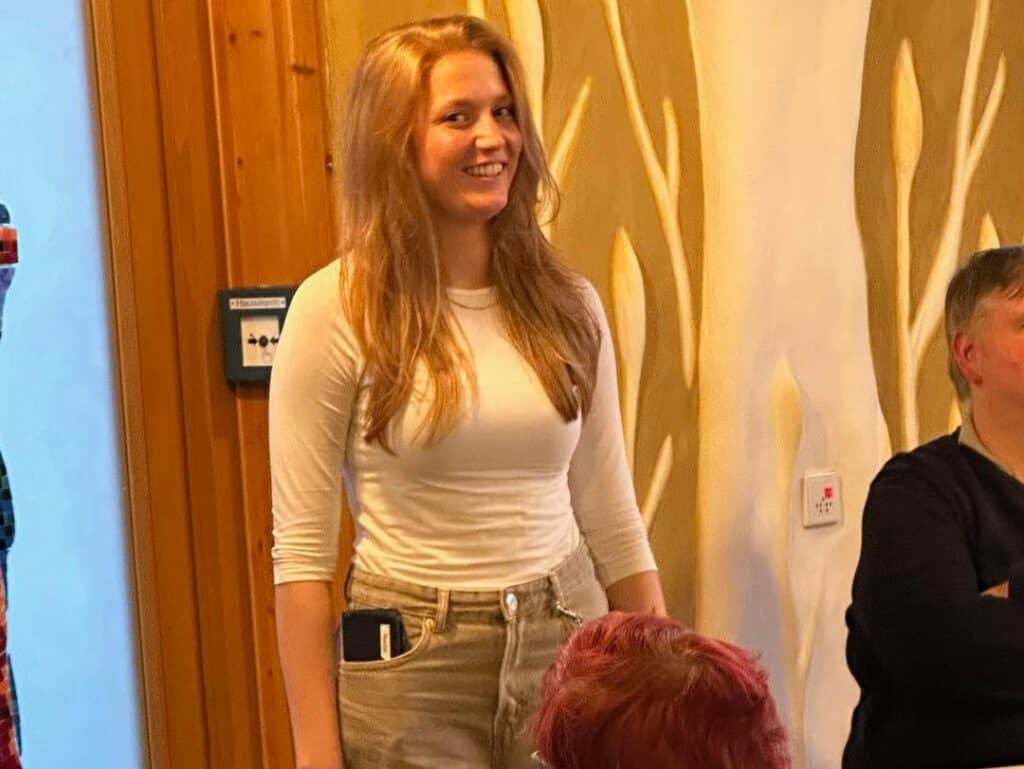 Eine Frau in einem weißen Hemd steht und lächelt in einem Raum mit Holzwänden und sitzenden Menschen in der Nähe.