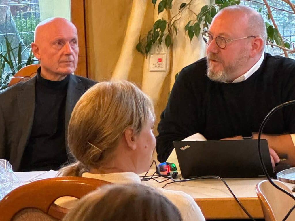 Zwei Männer sitzen an einem Tisch mit einem Laptop und sprechen mit einer Frau im Vordergrund.
