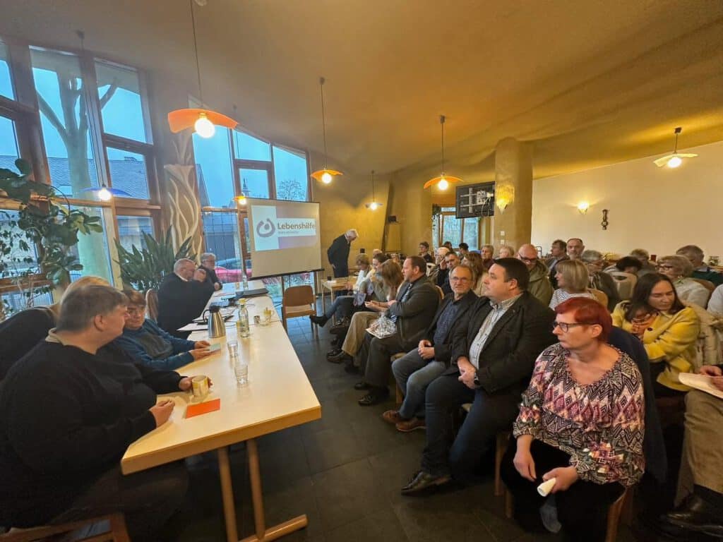 Menschen sitzen in einem Raum vor einer Tafel und einem Bildschirm, auf dem ein Logo der Lebenshilfe zu sehen ist.