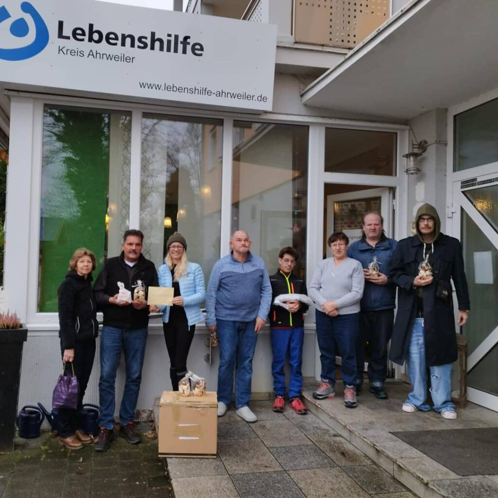 Acht Personen stehen vor einem Gebäude und halten Gegenstände und eine Kiste mit der Aufschrift Lebenshilfe.