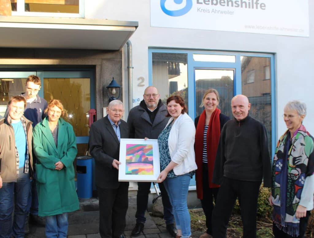 Eine Gruppe von Menschen steht vor einem Gebäude und hält ein bunt gerahmtes Kunstwerk in der Hand.
