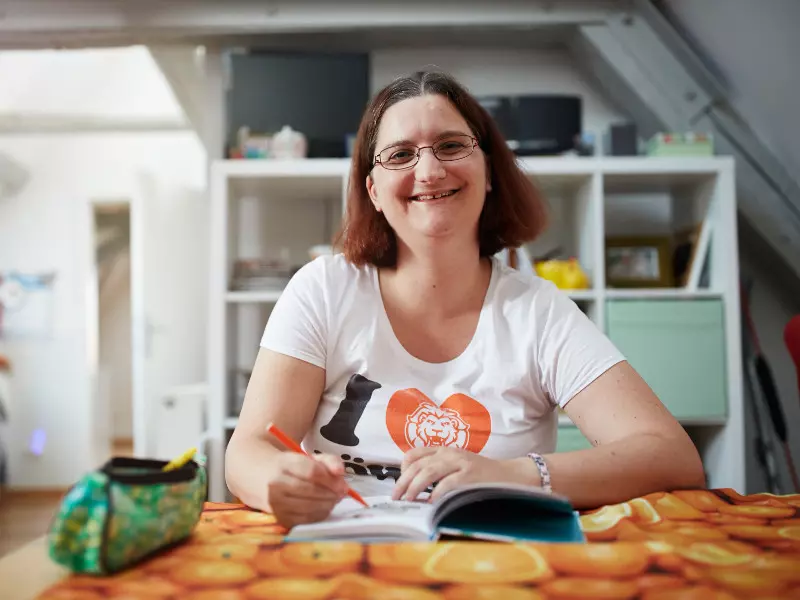 Eine Frau mit Brille und weißem T-Shirt sitzt lächelnd an einem Tisch und schreibt mit einem roten Stift in ein Notizbuch. Im Hintergrund sind ein Federmäppchen und Regale mit verschiedenen Gegenständen zu sehen.
