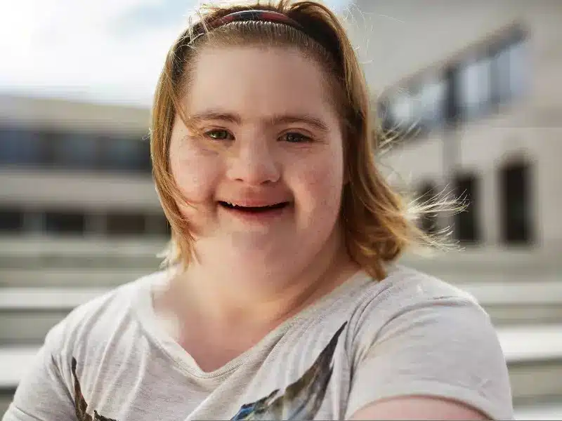 Eine junge Frau mit Down-Syndrom lächelt herzlich, während sie an einem sonnigen Tag im Freien sitzt, mit verschwommenen Gebäuden im Hintergrund.