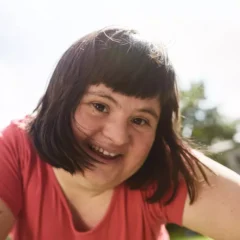 Eine Frau mit Down-Syndrom und dunklem Haar lächelt an einem sonnigen Tag im Freien und trägt ein rosa Hemd. Der Hintergrund ist verschwommen, Bäume und ein Gebäude sind sichtbar.