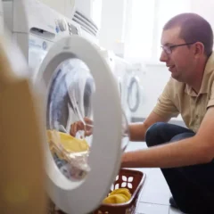 Ein Mann mit Brille und beigem Poloshirt kniet vor einer Frontlader-Waschmaschine und legt gelbe Wäschestücke hinein. Neben ihm steht ein brauner Wäschekorb mit weiterer gelber Wäsche auf dem Boden.