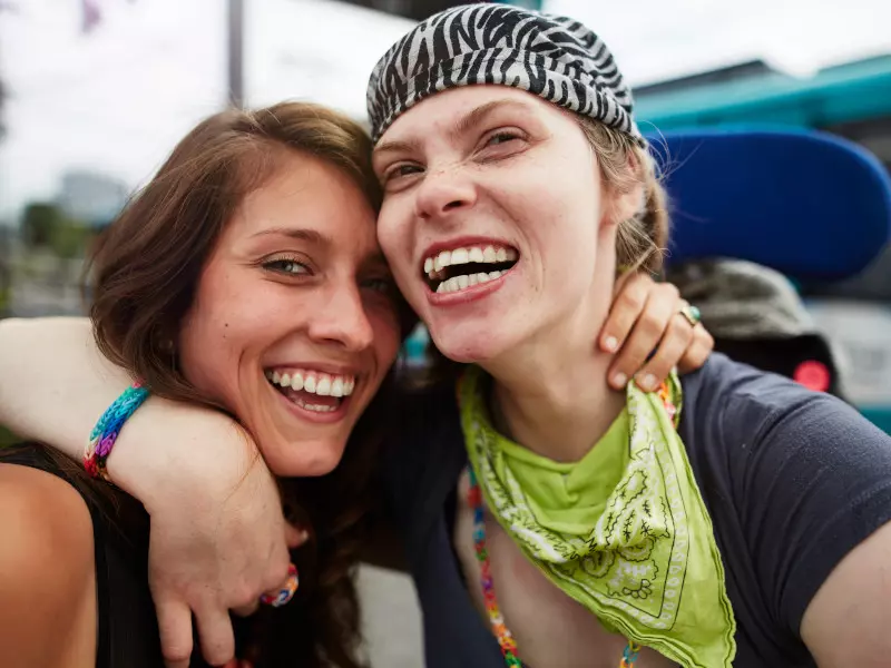 Zwei lächelnde Freunde posieren für ein Selfie im Freien. Einer hat den Arm um den anderen gelegt und trägt ein Stirnband mit Zebramuster und ein grünes Bandana. Beide sehen glücklich und fröhlich aus.
