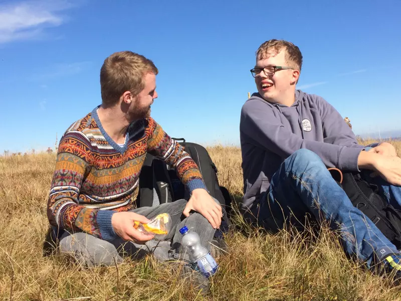 Zwei Menschen sitzen unter einem klaren, blauen Himmel im trockenen Gras, unterhalten sich und lächeln. Einer hält ein Sandwich und eine Wasserflasche in der Hand und trägt einen gemusterten Pullover; der andere trägt eine Brille und einen grauen Kapuzenpullover. Neben ihnen stehen Rucksäcke.