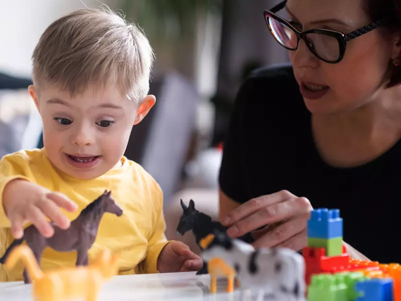 Ein kleiner Junge mit Down-Syndrom greift aufgeregt nach Spielzeugtieren auf einem Tisch, während eine erwachsene Frau mit Brille neben ihm sitzt und ihm beim Spielen und mit bunten Bauklötzen hilft.