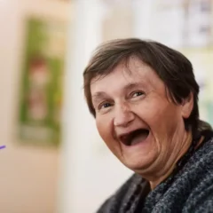 Eine ältere Frau mit kurzen Haaren und grauem Pullover sitzt fröhlich lächelnd im Haus. Der Hintergrund ist leicht verschwommen, im Vordergrund ist ein Getränk mit einem lila Strohhalm zu sehen.