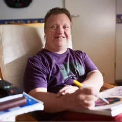 Ein Mann mit Down-Syndrom sitzt lächelnd in einem gemütlichen, hellen Raum auf einem Stuhl und zeichnet mit Buntstiften auf Papier. Auf dem Tisch vor ihm liegen Malutensilien und bunte Zeichnungen.
