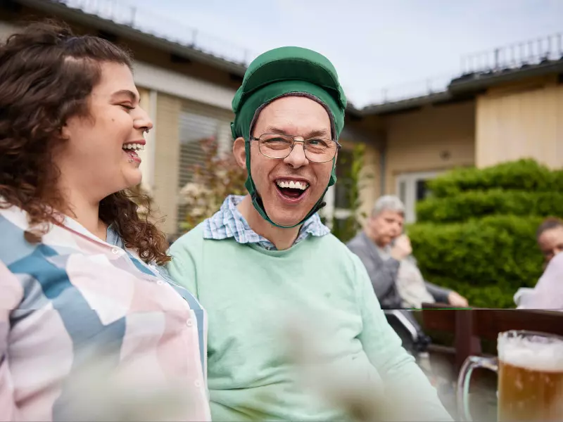 Ein Mann mit grünem Schutzhelm und Brille sitzt mit einem breiten Lächeln im Freien neben einer lachenden Frau. Im Hintergrund sitzen weitere Personen an einem Tisch in einer Gartenanlage.