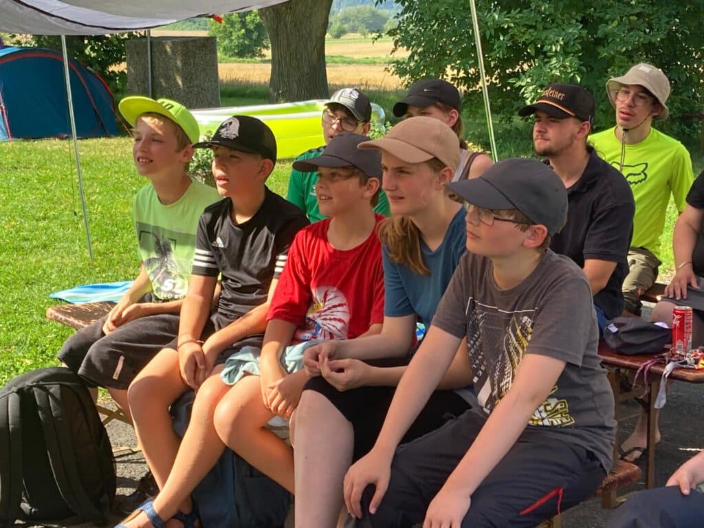 Eine Gruppe von Kindern und Jugendlichen sitzt im Freien auf Bänken unter einem Vordach und beobachtet etwas.
