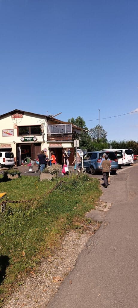 Menschen versammeln sich vor einem kleinen Gebäude am Straßenrand, in dessen Nähe Autos geparkt sind, und der Himmel ist klar.
