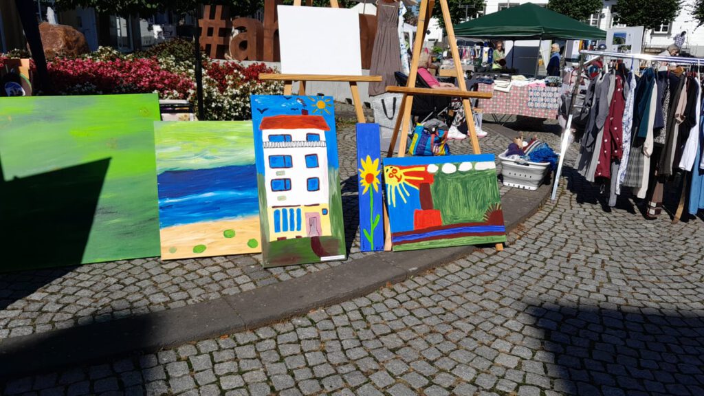 Farbenfrohe Gemälde, die auf Staffeleien und auf dem Boden auf einem Markt mit Kleiderständen ausgestellt sind.