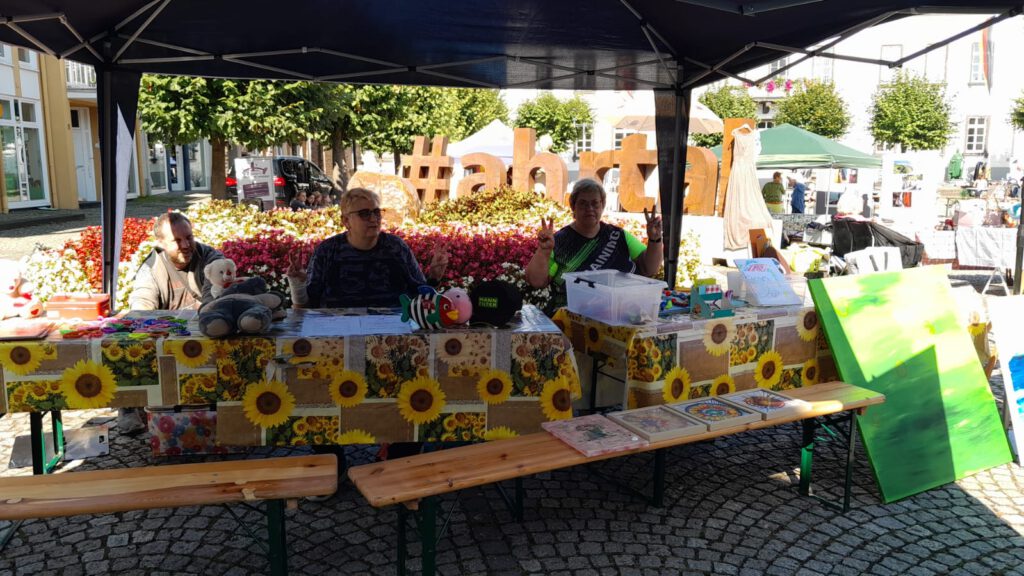 Drei Personen sitzen an einem mit Sonnenblumen geschmückten Stand auf einem Markt unter einem Vordach.