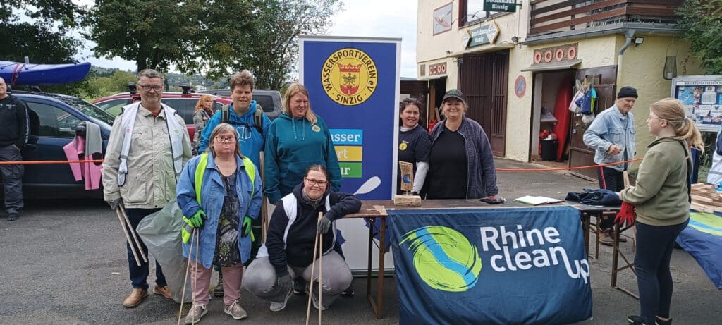 Eine Gruppe von Menschen posiert bei einer Rhein-Säuberungsaktion im Freien mit Bannern und Reinigungsgeräten.