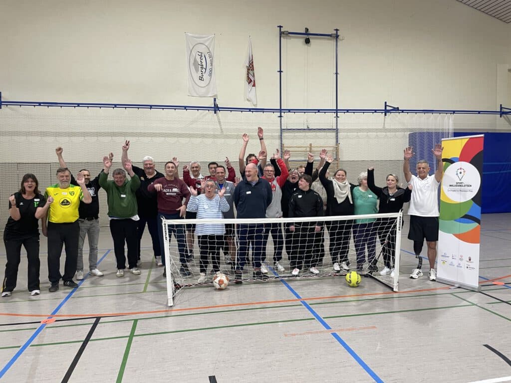 Eine Gruppe von Erwachsenen posiert mit erhobenen Armen hinter einem Fußballnetz in einer Turnhalle.