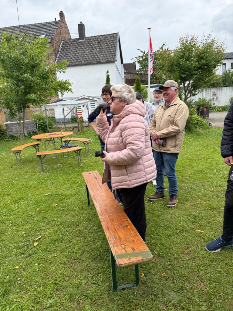 Mehrere Personen stehen in der Nähe von Holzbänken auf einer Wiese mit Bäumen und Häusern im Hintergrund.