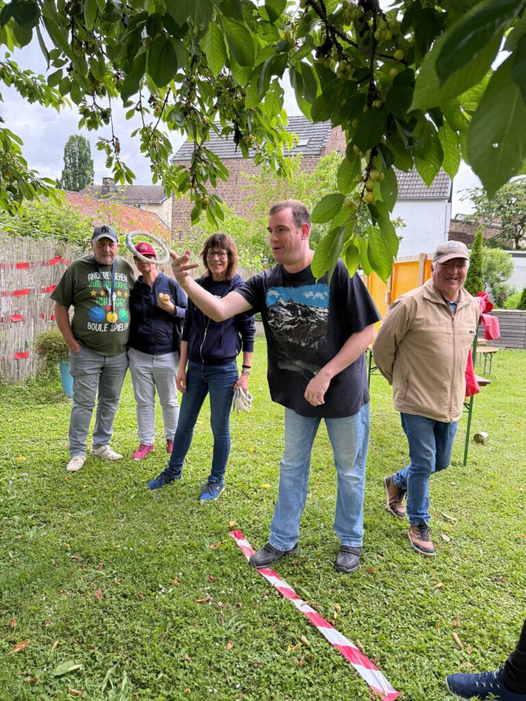 Fünf Personen stehen auf einer Wiese; ein Mann wirft bei einem Spiel im Freien einen Ball unter einen Baum.