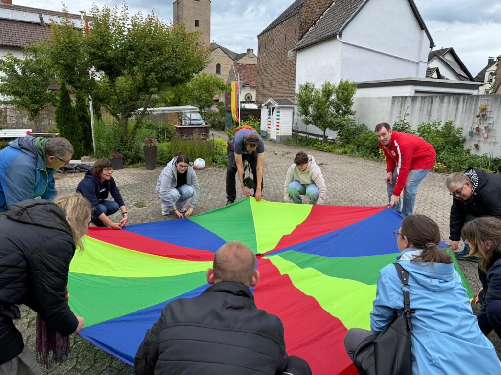 Eine Gruppe von Menschen hält und spannt einen bunten Fallschirm in einem gepflasterten Innenhof.