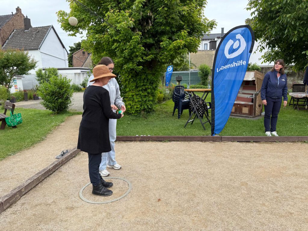 Zwei Personen stehen auf einem Pétanque-Platz, in der Nähe ein Banner der Lebenshilfe und andere im Hintergrund.
