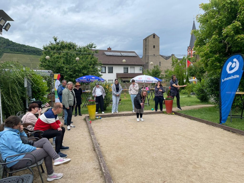 Eine Gruppe von Menschen spielt Pétanque im Freien, mit Zuschauern und Gebäuden im Hintergrund.