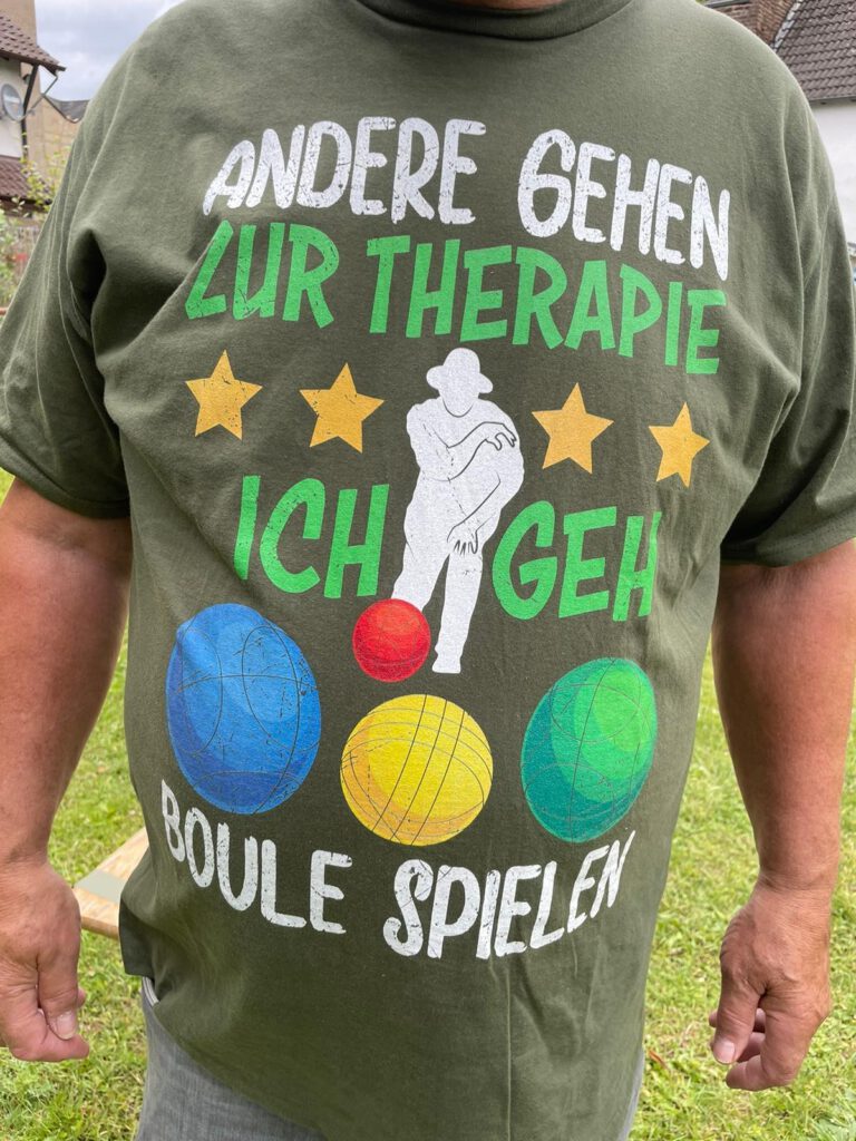 Eine Person trägt ein grünes T-Shirt mit der Aufschrift "Andere gehen zur Therapie, ich geh Boule spielen".