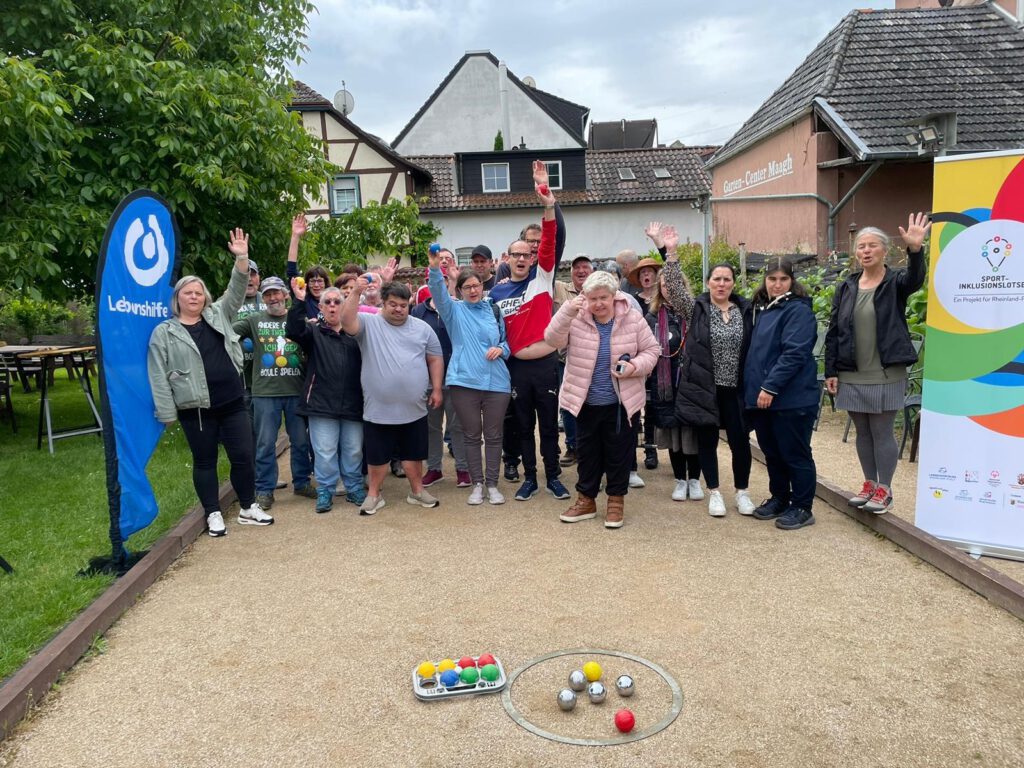 Eine Gruppe von Menschen posiert und winkt neben Bocciakugeln auf einem Platz im Freien.