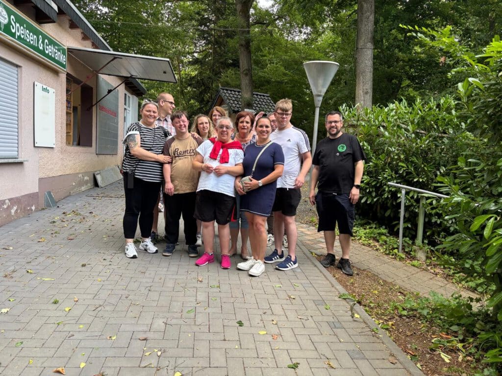 Eine Gruppe von elf Personen steht gemeinsam auf einem gepflasterten Weg vor einem Gebäude in der Nähe einer Grünanlage.