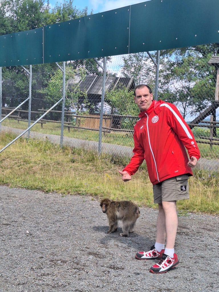 Ein Mann in einer roten Jacke zeigt auf einen kleinen Hund, der auf Kies neben einem Maschendrahtzaun steht.