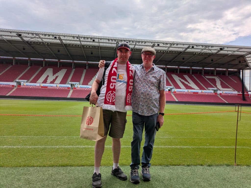 Zwei Männer stehen auf einem Fußballplatz, im Hintergrund sind Stadionränge und der Schriftzug „MAINZ“ zu sehen.