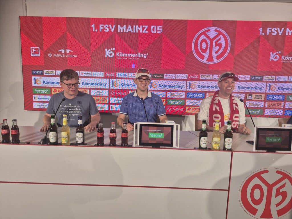 Drei Personen sitzen an einem Pressekonferenztisch mit dem Logo von Mainz 05 und verschiedenen Getränken.